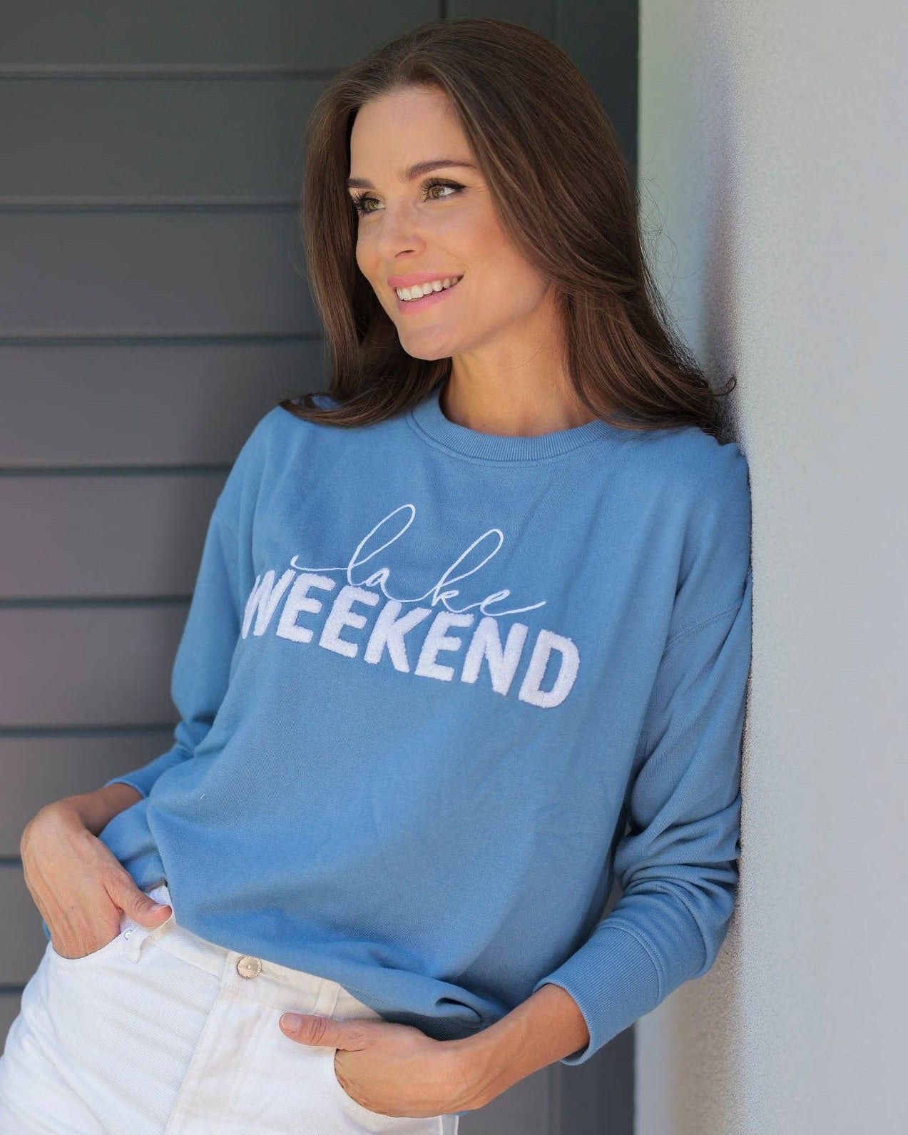 Woman wearing a blue sweatshirt with text, standing against a gray wall.